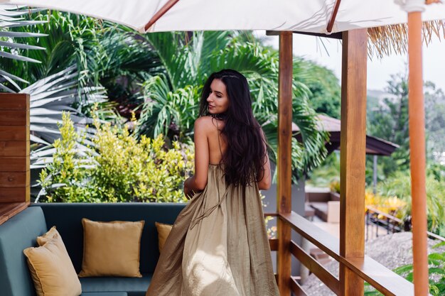 Donna sorridente positiva abbastanza europea in vestito da volo estivo alla luce del giorno naturale sulla terrazza della villa godendo di una bella vacanza, divano all'aperto con cuscini su tropicale