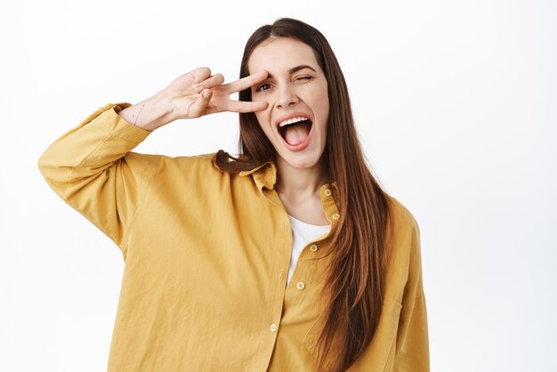 Donna sorridente positiva che mostra il gesto kawaii vsign e la bocca aperta ammiccante felice di stare sul lato positivo in piedi ottimista su sfondo bianco