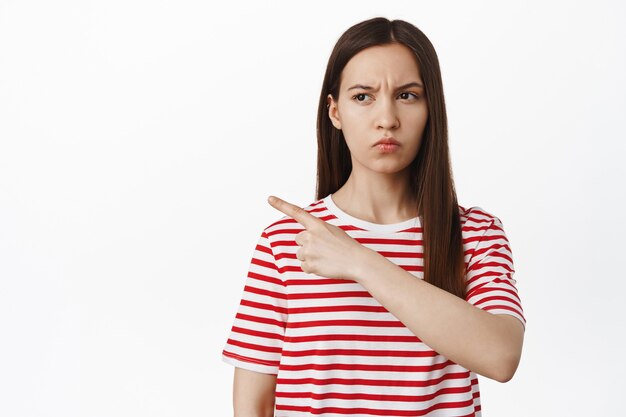 Donna sospettosa che strizza gli occhi, indica e guarda a sinistra con un viso dubbioso, incredulo, in piedi in maglietta contro il muro bianco.