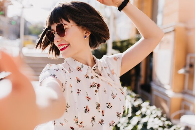 Donna spensierata in camicetta elegante che balla per strada. Modello femminile di risata con capelli castano scuro corti che fa selfie sulla città di sfocatura.