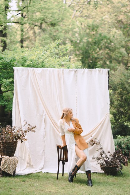 Donna splendida in vestito bianco e rivestimento marrone che stanno e che pensano nel giardino con la parete bianca durante il giorno.