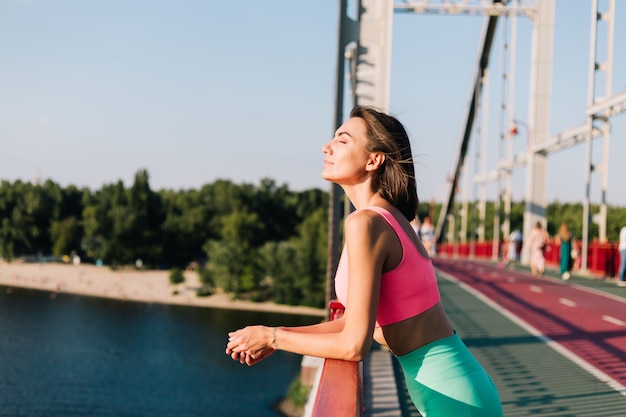 Donna sportiva che indossa abbigliamento sportivo al tramonto sul moderno ponte con vista sul fiume, guardati intorno godendoti il clima estivo