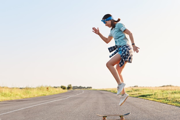 Donna sportiva che indossa t-shirt e short facendo acrobazie su uno skateboard su strada su strada asfaltata, saltando in aria, godendosi lo skateboard da solo al tramonto in estate.