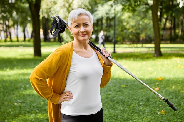 Donna sportiva dai capelli corti in pensione che dice di sì a uno stile di vita sano e attivo, tiene il bastone per il nordic walking sulle spalle, farà una bella passeggiata, allena il corpo e il sistema cardiovascolare