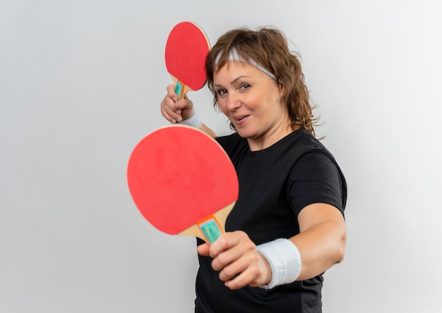 Donna sportiva di mezza età in maglietta nera con archetto che tiene due racchette per ping-pong con il sorriso sul viso in piedi sopra il muro bianco