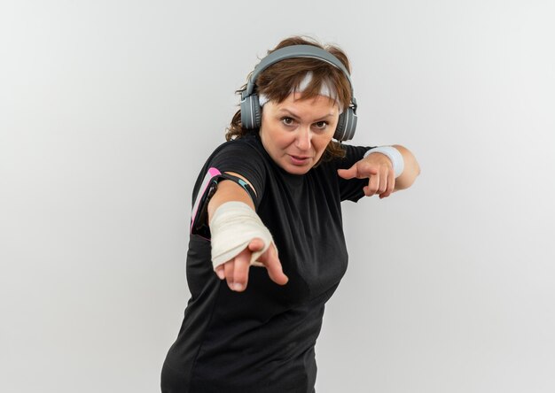 Donna sportiva di mezza età in maglietta nera con archetto e cuffie che puntano con le dita indice in piedi sul muro bianco