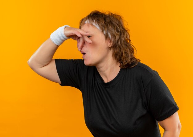 Donna sportiva di mezza età in maglietta nera con fascia che chiude il naso con le dita che soffrono di puzza in piedi sopra la parete arancione