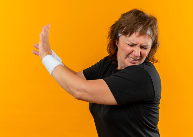 Donna sportiva di mezza età in maglietta nera con fascia che fa gesto di difesa con le mani in piedi sopra la parete arancione