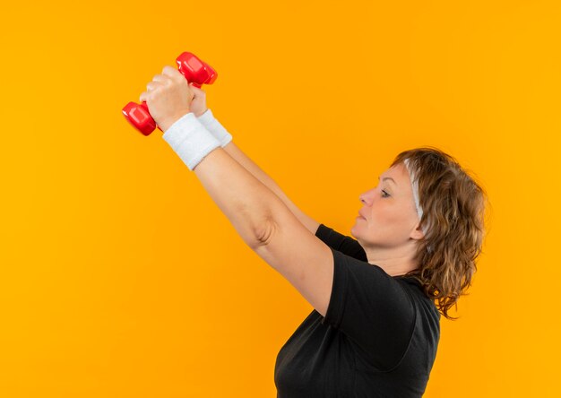 Donna sportiva di mezza età in maglietta nera con fascia che lavora con manubri alzando le mani guardando fiducioso e teso in piedi sopra la parete arancione