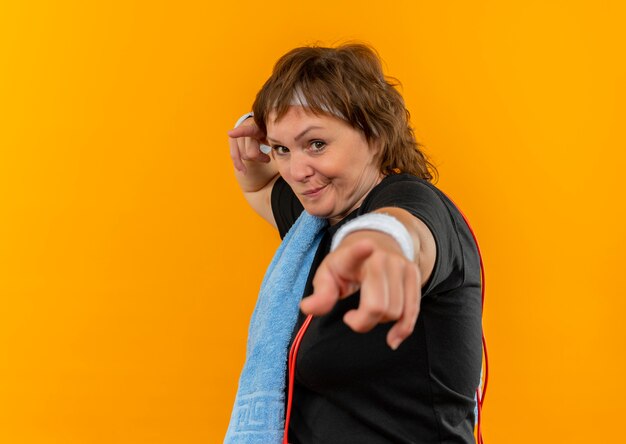 Donna sportiva di mezza età in maglietta nera con fascia e con asciugamano sulla spalla che punta con le dita che guardano con espressione scettica in piedi sopra la parete arancione