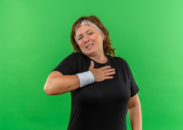 Donna sportiva di mezza età in maglietta nera con la fascia che tiene la mano sul petto alla ricerca di malessere in piedi sul muro verde