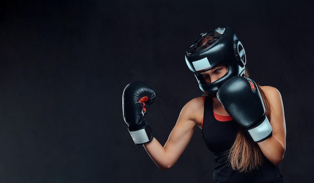 Donna sportiva in abbigliamento sportivo che indossa un casco protettivo e guanti da boxe, allenamento in palestra. Isolato su sfondo scuro con texture.