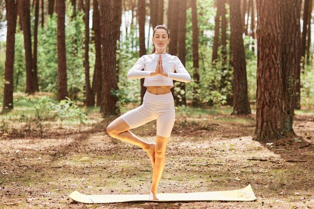 Donna sportiva rilassata in piedi all'aria aperta con gli occhi chiusi tenendo i palmi uniti, in piedi su una gamba sola, indossando abbigliamento sportivo, godendosi l'allenamento nella bellissima foresta.