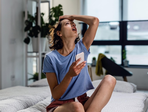 Donna stressata che riceve un messaggio di testo sul suo telefono e guarda incredula mentre è seduta sul letto