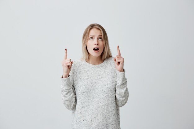 Donna stupita con i capelli tinti di biondo, in maglione che guarda e punta verso l'alto con le dita sorprese nel vedere qualcosa. Giovane donna emozionata interessata con la bocca spalancata