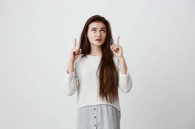 Donna stupita con lunghi capelli scuri dritti, indossando abiti casual, guardando con gli occhi scuri verso l'alto e la bocca ampiamente aperta, indicando con gli indici lo spazio della copia