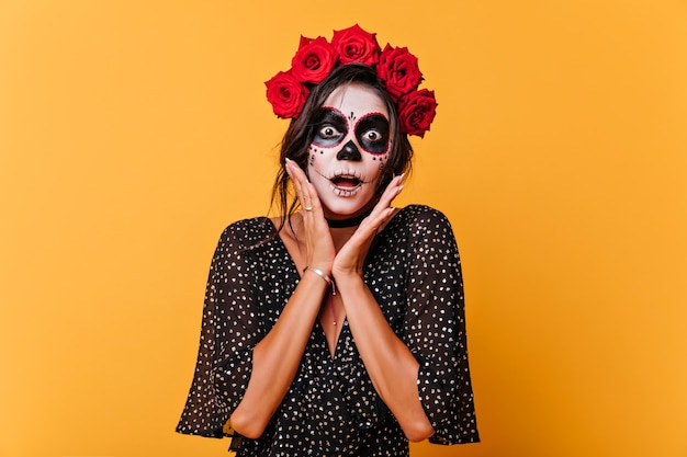 Donna stupita con rose rosse nei capelli che celebra halloween. Ragazza spaventosa con trucco muertos in posa su sfondo giallo.