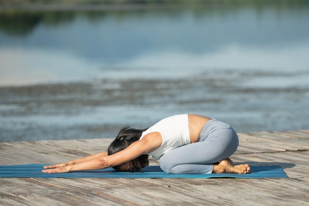 Donna su una stuoia di yoga per rilassarsi nel parco al lago di montagna. attraente ragazza sportiva in abiti sportivi. ragazza sportiva che lavora fuori. Stile di vita sportivo sano. Giovane donna atletica nel fare esercizio di fitness.