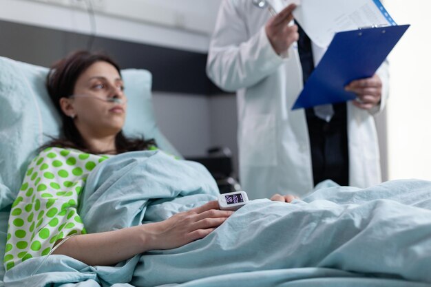 Donna sul letto d'ospedale con pulsossimetro sul dito che mostra una bassa saturazione di ossigeno ascoltando i risultati del laboratorio di lettura del medico dagli appunti. Paziente con cannula nasale che riceve i risultati della diagnosi.