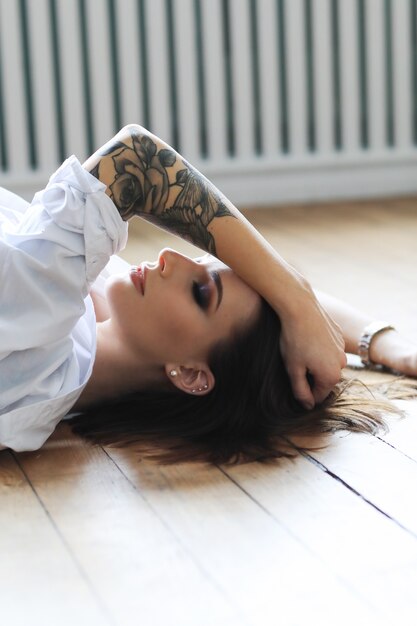 Donna tatuata in una camicia bianca che si trova sul pavimento di legno