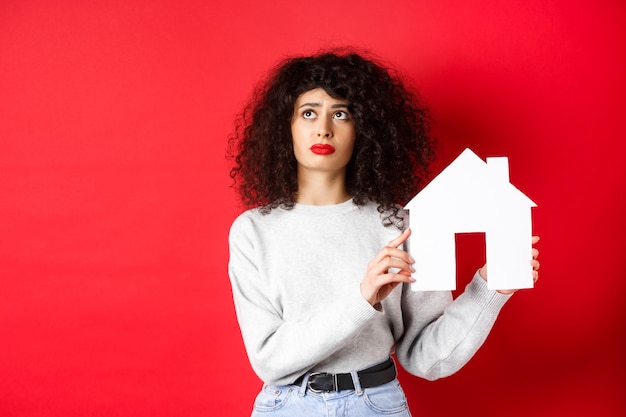 Donna triste immobiliare che pensa di acquistare la propria casa mostrando il ritaglio di carta e guardando in alto con usp...