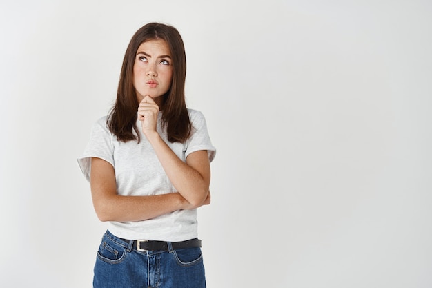 Donna turbata che pensa alla soluzione, labbra increspate pensose e guardando in alto, facendo una scelta o una decisione, in piedi sul muro bianco