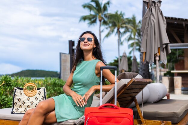 Donna turistica elegante caucasica capelli abbastanza lunghi felice in vestito con la valigia rossa fuori dall'hotel