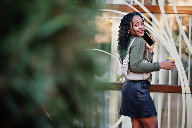 Donne afroamericane alla moda alla moda in maglione verde e gonna nera poste in un caffè all'aperto con una tazza di caffè e parlare sul telefono cellulare