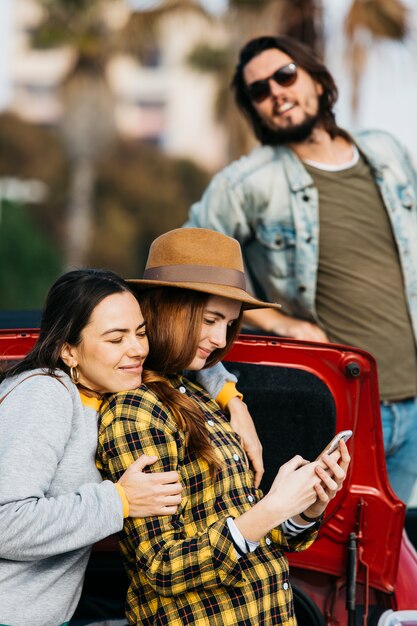 Donne allegre che abbracciano signora con smartphone vicino a tronco d&#39;auto e l&#39;uomo che si appoggia fuori dall&#39;automobile