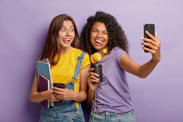 Donne allegre studiano in un gruppo, si divertono durante la pausa al college, prendono selfie sullo smartphone, mostrano le lingue, tengono bicchieri di carta di caffè, tengono blocchi per appunti, posano insieme contro il muro viola.