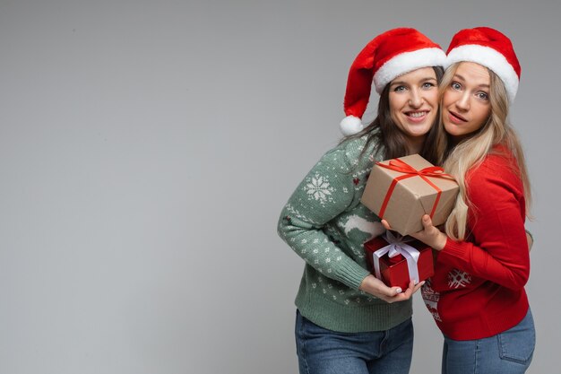 Donne attraenti amiche con cappelli natalizi rossi e bianchi si tengono regali l'una per l'altra e sorridono