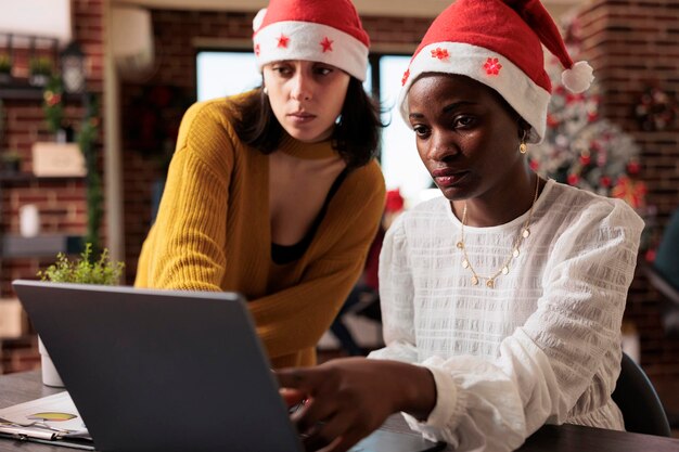 Donne che fanno lavoro di squadra in un ufficio festivo, lavorando su un laptop per pianificare il progetto in uno spazio pieno di ornamenti e luci natalizie. Persone al lavoro d'ufficio con albero di Natale e decorazioni.