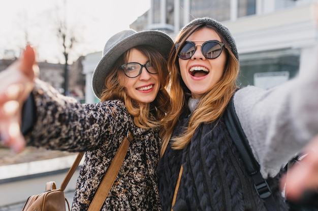 Donne eccitate in occhiali alla moda che si divertono durante la passeggiata mattutina per la città. Ritratto all'aperto di due amici allegri in cappelli alla moda che fanno selfie e ridendo, agitando le mani.