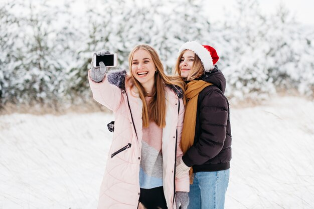 Donne felici in cappello di Santa prendendo selfie nella foresta di inverno
