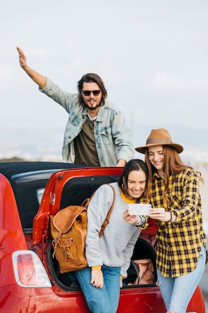 Donne positive che prendono selfie sullo smartphone vicino al tronco di automobile e all&#39;uomo che si appoggia fuori dall&#39;auto