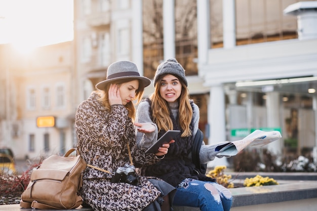 Donne sorridenti alla moda che si siedono nel centro della città che esprimono emozioni luminose in una giornata di sole in città. Buon viaggio insieme, cercando di trovare la posizione sulla mappa.