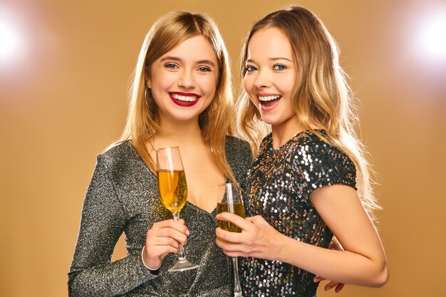 Donne sorridenti felici in eleganti abiti glamour con bicchieri di champagne sul muro d'oro