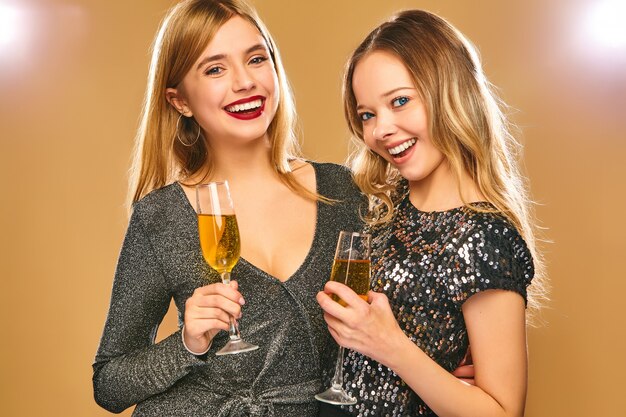 Donne sorridenti felici in eleganti abiti glamour con bicchieri di champagne sul muro d'oro