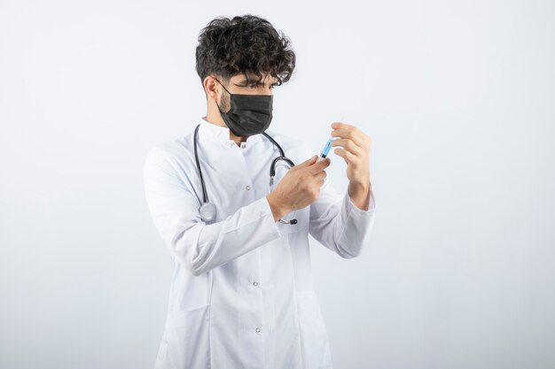 Dottore in camice bianco con stetoscopio che tiene e tocca una siringa isolata su bianco.