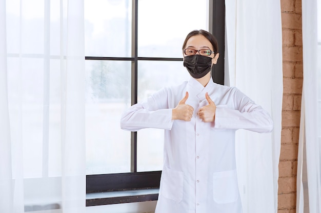 Dottore in maschera nera e occhiali in piedi vicino alla finestra e mostrando segno di divertimento.