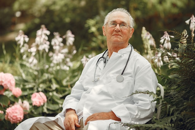 Dottore in uniforme bianca. Il vecchio uomo seduto in un parco estivo. Anziano con lo stetoscopio.