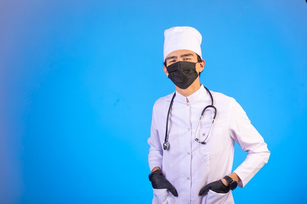 Dottore in uniforme medica bianca con stetoscopio, maschera facciale e mani in tasca.