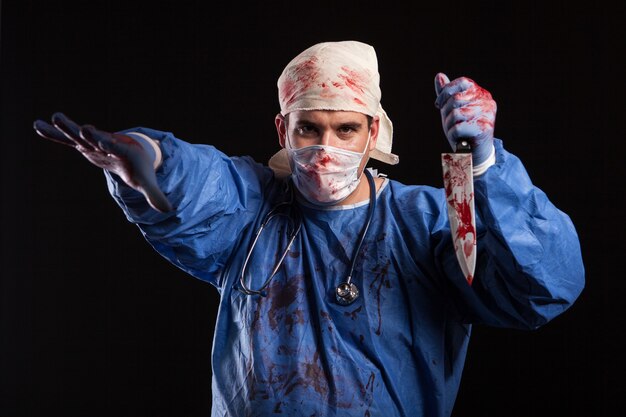 Dottore pazzo che tiene un coltello coperto di sangue in studio su sfondo nero. Medico maniaco con maschera sul viso per halloween.