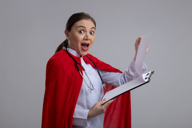 Dottoressa con stetoscopio in uniforme medica bianca e mantello da supereroe rosso che mostra appunti con pagine vuote guardando davanti stupito e preoccupato in piedi sopra il muro bianco