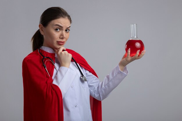 Dottoressa con stetoscopio in uniforme medica bianca e mantello da supereroe rosso che tiene la boccetta con liquido rosso guardando davanti con espressione pensierosa pensando in piedi sopra il muro bianco