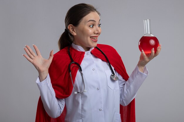 Dottoressa con stetoscopio in uniforme medica bianca e mantello da supereroe rosso che tiene la boccetta con liquido rosso guardandolo in piedi felice ed eccitato sopra il muro bianco