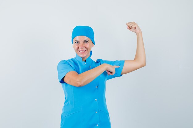 Dottoressa in uniforme blu che punta i muscoli del braccio e guardando fiducioso, vista frontale.