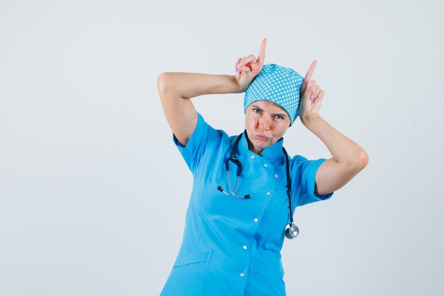 Dottoressa in uniforme blu tenendo le dita sopra la testa come corna di toro e guardando divertente, vista frontale.