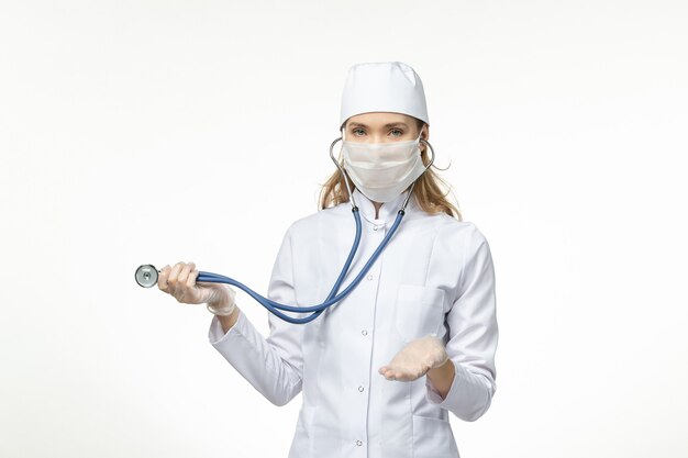 Dottoressa vista frontale in tuta medica bianca con maschera sterile a causa del coronavirus utilizzando uno stetoscopio sulla scrivania bianca malattia covid- malattia pandemica