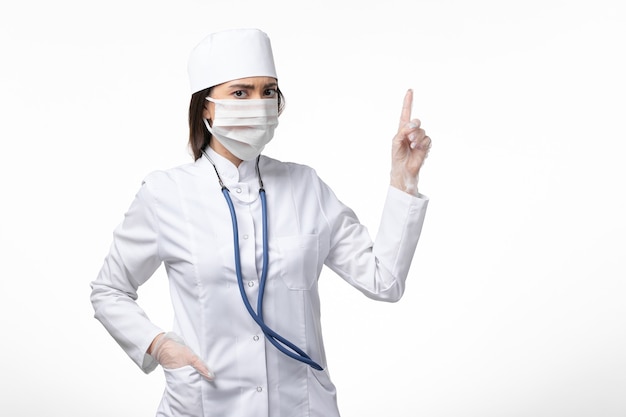 Dottoressa vista frontale in tuta medica bianca sterile con maschera a causa del coronavirus che alza il dito sul muro bianco malattia malattia virus pandemico covid-